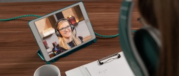 OnlineCoaching in der faw Blick über die Schulter einer Person auf ein Tablet auf einem Tisch; auf dem Bildschirm eine blonde Frau mit Headset