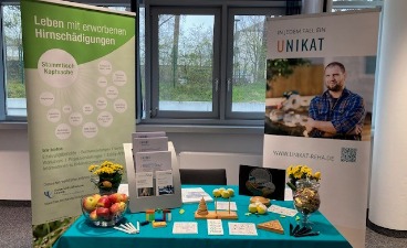 Die faw auf dem 16. Nachsorgekongress Foto: Kongressstand der faw auf dem 16. Nachsorgekongress: Im Vordergrund der faw-Messestand samt Infomaterial, Stiften,, aber auch Äpfeln und Blumen. Dahinter zwei Roll-Ups, eines zum Thema Leben mit erworbenen Hirnschädigungen und eines zum Thema UNIKAT.