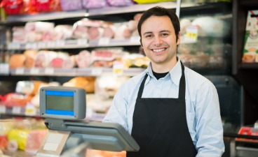 TQplus Verkäufer*in Beispielfoto: Beispielfoto: Person in schwarzer Schürze hinter einer Theke in einem Lebensmittelgeschäft. Im Hintergrund Regale mit Feinkostprodukten.
