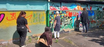 Drei Menschen stehen, eine sitzt vor einer Mauer. Die beiden rechts stehenden Menschen (ein Mann, eine Frau) sprühen gerade Muster auf die Mauer. Die beiden Mädchen links unterhalten sich.