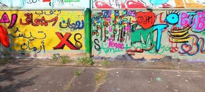 Eine bunte Graffiti Wand. Linke Seite ist gelb mit einem roten X und anderen Zeichen, wahrscheinlich arabisch. Rechte Seite bunt mit Buchstaben in Grün und Türkis und einem riten herzen mit den Buchstaben DJM