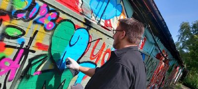 Das Bild zeigt einen Auschnitt einer Graffiti Mauer von unten nach oben fotografiert. Ein junger Mann spüht gerade ein Herz in grün und türkis auf die Mauer.