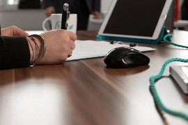 Foto: Nahaufnahme einer Hand mit Stift am Schreibtisch, die sich Notizen macht.