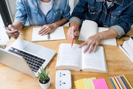 Foto: Blick auf einen Schreibtisch mit Laptop, an dem zwei Menschen zusammen lernen.