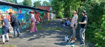 Das Bild zeigt eine kleine Straße oder Weg, der von einer mit Graffitis übersäten Mauer begrenzt ist. Davor stehen eine Frau, ein junger Mann und eine junge Frau, die gerade Farbe auf die mauer sprühen. Am rechten Straßenrand stehen zwei Jugendliche - vor ihnen stehen die Spraymaterialien, weiter hinten sitzen zwei Jugendliche