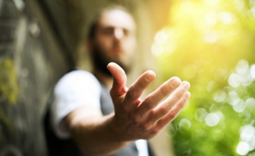 Foto: Eine Person streckt die Hand aus, als ob sie etwas anbieten oder empfangen möchte. Der Hintergrund ist unscharf mit Sonnenlicht und Bäumen.