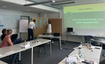 Schulungsraum, in dem mehrere Menschen an Tischen zum Referenten am Whiteboard sehen.