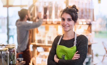 Verkäuferin mit grüner Arbeitsschürze steht mit verschränkten Armen im Laden, im Hintergrund räumt ein Kollege Waren in ein Regal.