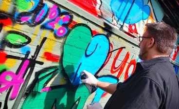 Zu sehen ist ein Mann im grauen Hemd, der im Sonnenschein ein Graffiti in grün und blau an eine bereits mit anderen Graffitis verzierten Wand sprüht.