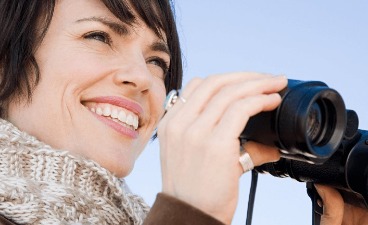 Auf dem Bild ist eine Frau zusehen, die lächelnd in die Ferne sieht und dabei ein Fernglas in den Händen hält.