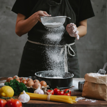 Workshop: Rezept für den Wiedereinsteig Beispielfoto Küche: Mehl wird über eine Backform gesiebt, Auf dem Arbeitstisch Kochzutaten, wie Pasta, Eier, Tomaten, etc.