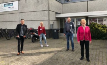 Foto: Vier Menschen stehen im Abstand zueinander vor einem Gebäude. 