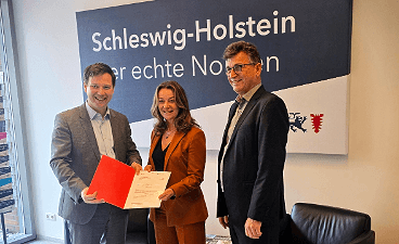 Foto: Staatssekretär u. zwei Mitarbeitende der faw bei der Dokumentenübergabe vor einem Schild "Schleswig-Holstein Der echte Norden".