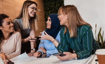 Foto: Vier junge Frauen unterschiedlicher ethnischer Herkunft unterhalten sich fröhlich miteinander.