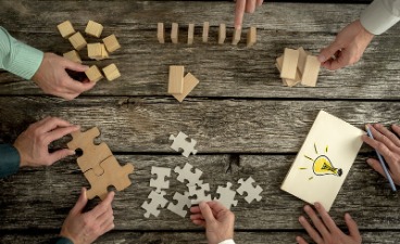 Beispielbild: Vogelperspektive: Auf einem Holztisch sind vier Handpaare zu sehen, zwei legen Puzzleteile, zwei Holzklötzchen und einer eine Holztafel auf der eine Glühbirne gezeichnet ist. 