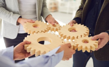 Symbolfoto für Team-/ Zusammenarbeit und das Ineinandergreifen verschiedener Prozesse: Drei Personen halten hölzerne, ineinandergreifende Zahnräder in den Händen.